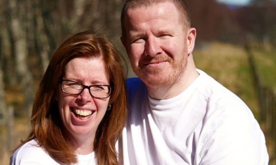 Susan and colin, a couple in their thirties, stand outside and smile at the camera.