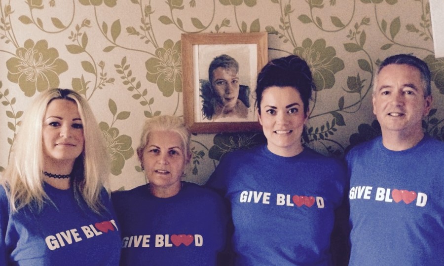 Lizzie stands with three members of Jordan's family (two young women and an older man, all wearing Give Blood t-shirts) in the living room. There is a portrait of Jordan on the wall.