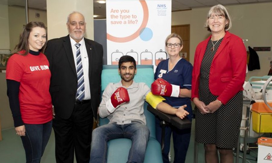 Boxer Kash Farooq sits on bed smiling while surrounded by members of the Scottish National Blood Transfusion Service and Scottish Ahlul Bayt Society. 