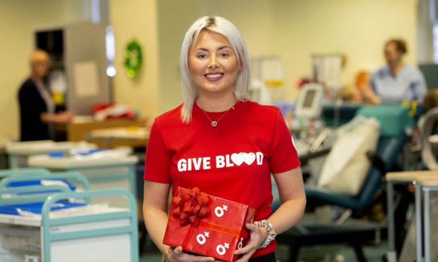 Jemma stands wearing a Give Blood t-shirt while holding a boxed Christmas present wrapped in O negative wrapping paper.  