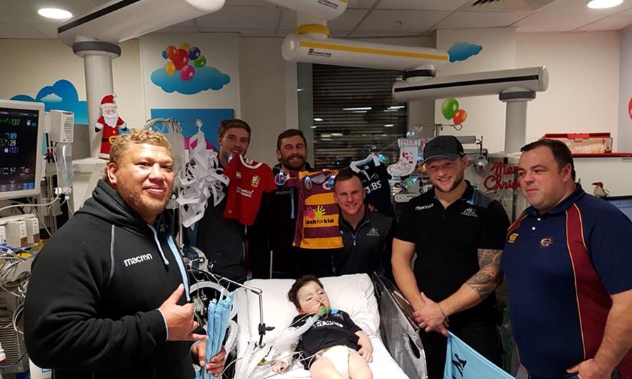 Members from rugby team Glasgow Warriors surround Iain while he lies in hospital bed. 
