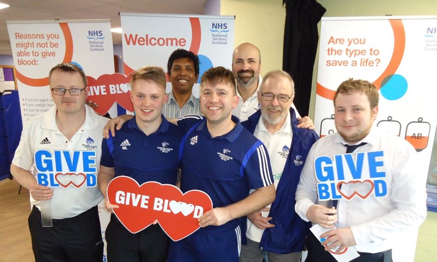 Members of Northern Counties Cricket Club smile and pose for photo while holding Give Blood signs. 