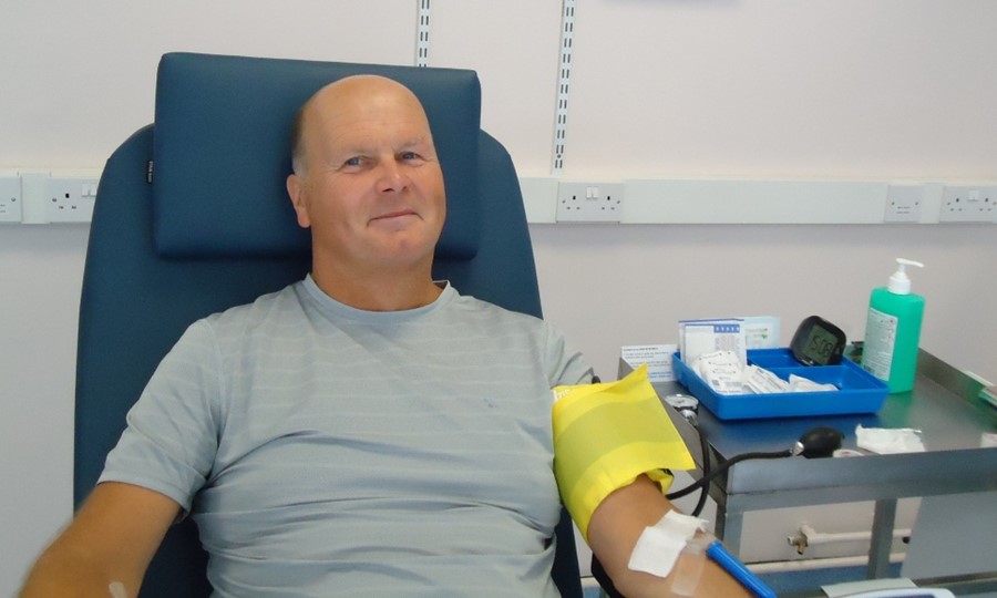 Paul sits on bed smiling after giving blood. 