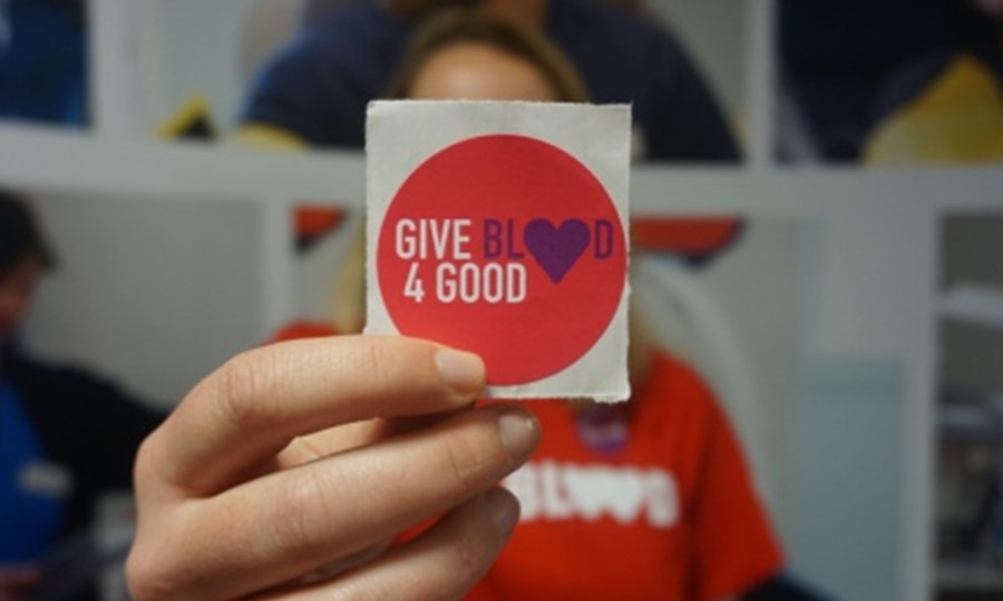 Young lady holding up a 'Give Blood 4 Good' sticker in front of her face