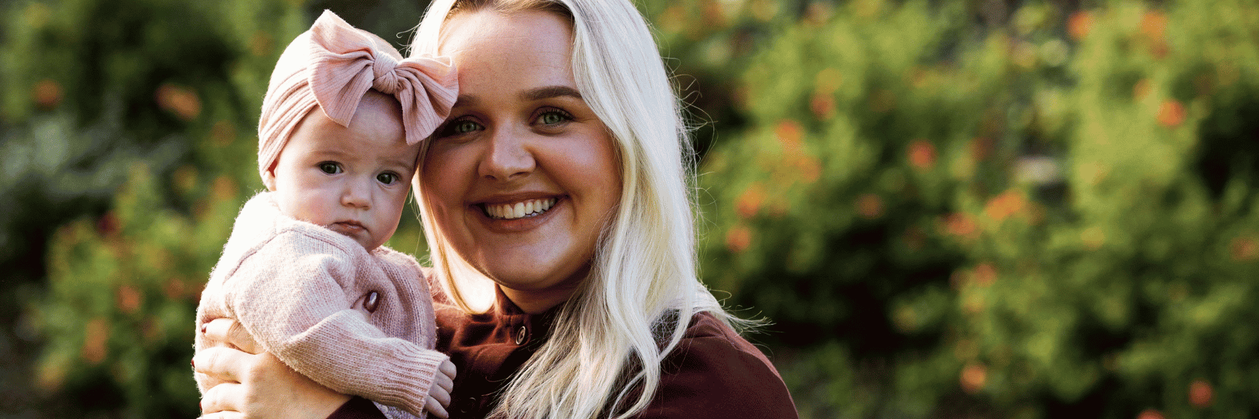 Blonde Mum Caitlyn poses with baby Millie outside against a leafy green backdrop