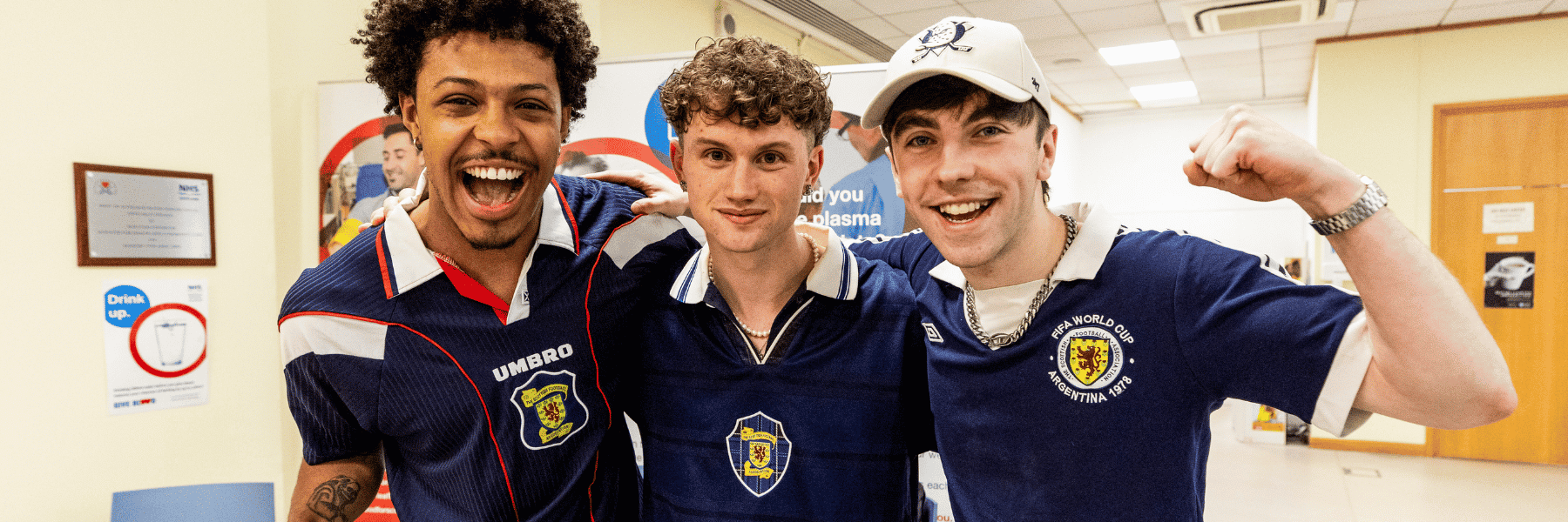 The three members of band Scratchcard Wednesday stand in Glasgow Blood Donor Centre wearing Scotland strips and smiling