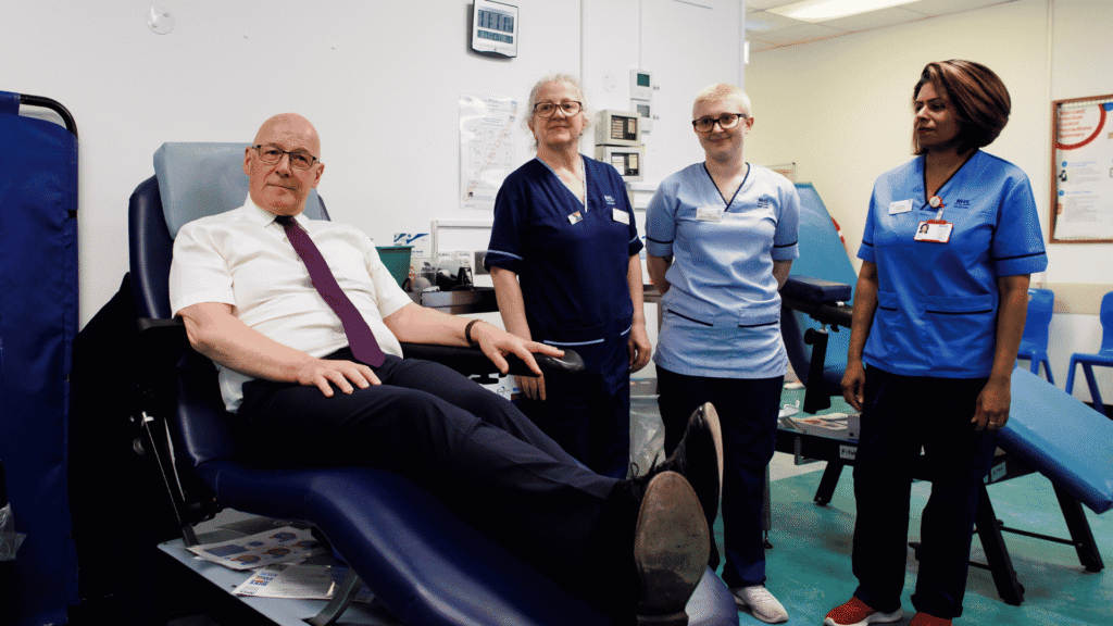 The First Minister is sitting on a bed about to donate blood, with three SNBTS members of staff standing by